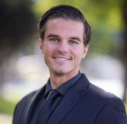Wood & Delgado | Professional headshot of a smiling male professional in a black suit and tie with a blurred outdoor background.